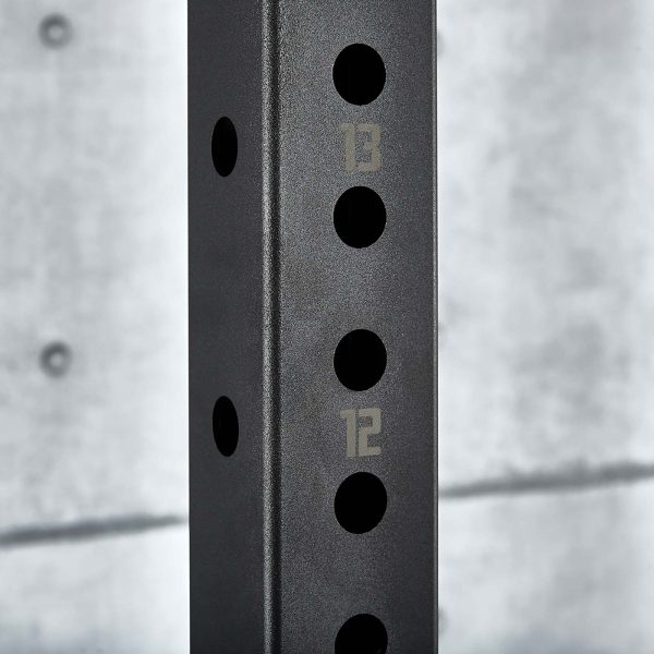 Close-up of a black metal post on the ATX® Power Rack 770 XL SET with numbered holes 12 and 13 visible against a textured, light gray concrete wall in the background.