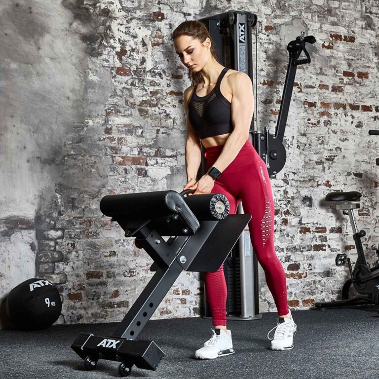 A woman in a black sports bra and red leggings uses gym equipment against a brick wall. She has light brown hair and is focused on adjusting the sissy squat stand. A slam ball and other gym equipment are visible in the background.