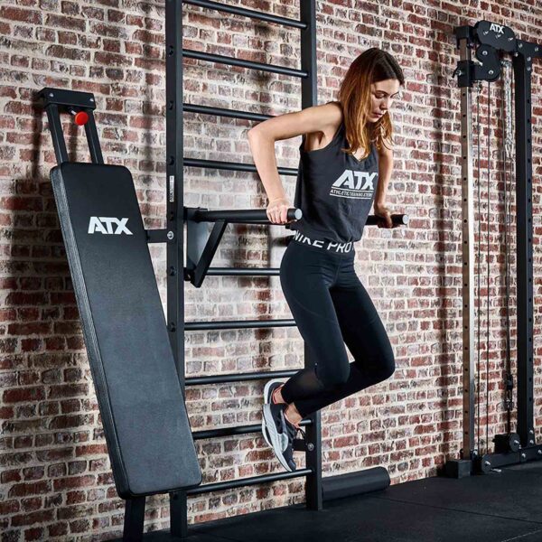 A person in workout attire performs tricep dips on parallel bars attached to the ATX® Wall Bar Gym, a wall-mounted exercise station with Swedish Bars, set in a gym with brick walls. Nearby is a padded incline bench.