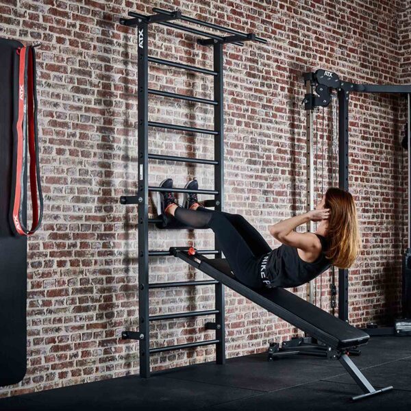 A person performs incline sit-ups on an ATX® Wall Bar Gym bench in a gym, located next to Swedish bars and other exercise equipment. Theyre wearing a black outfit and have their feet secured under a bar against the brick wall.