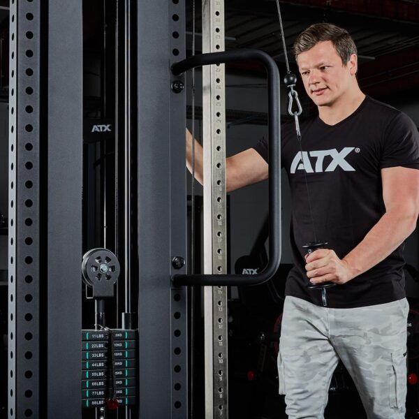 A man in a black ATX t-shirt and gray camo pants stands beside a cable machine in a gym, gripping the handle with ATX® Conic Fat Grips attached to the pulley system. The machine is loaded with weights.