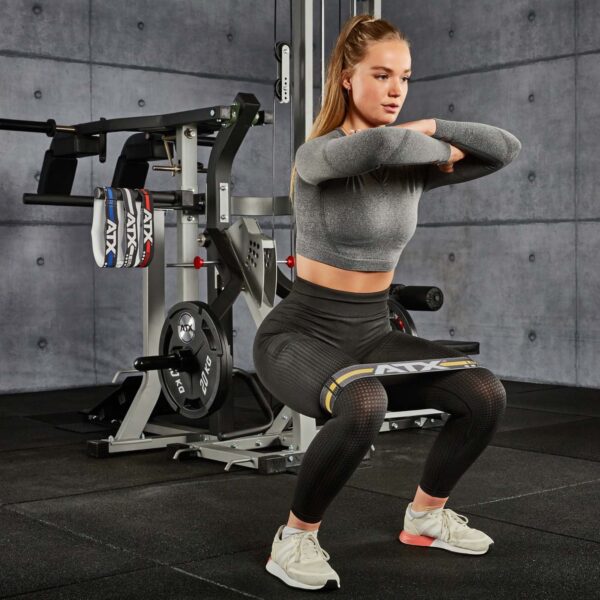 A woman in athletic attire performs a squat with an ATX® Loop Band positioned around her thighs in a gym setting. She maintains a focused expression, with exercise equipment visible in the background against a gray wall.