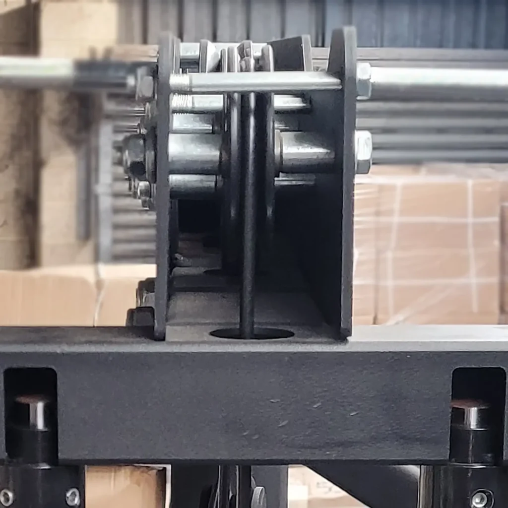 A close-up image of industrial machinery resembling a row machine, showcasing interconnected metal bolts, brackets, and pulleys. The background features cardboard boxes and a corrugated metal wall, partially visible. The focus is on the central pulley system.