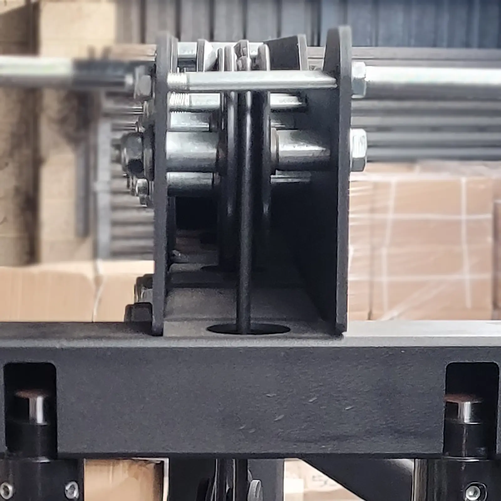 A close-up image of industrial machinery resembling a row machine, showcasing interconnected metal bolts, brackets, and pulleys. The background features cardboard boxes and a corrugated metal wall, partially visible. The focus is on the central pulley system.