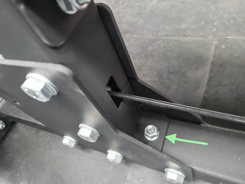Close-up of a mechanical hinge on a lat pulldown machine, with multiple bolts attached to a metal frame. A green arrow points to a specific bolt, highlighting its placement. The background consists of a dark tiled surface.