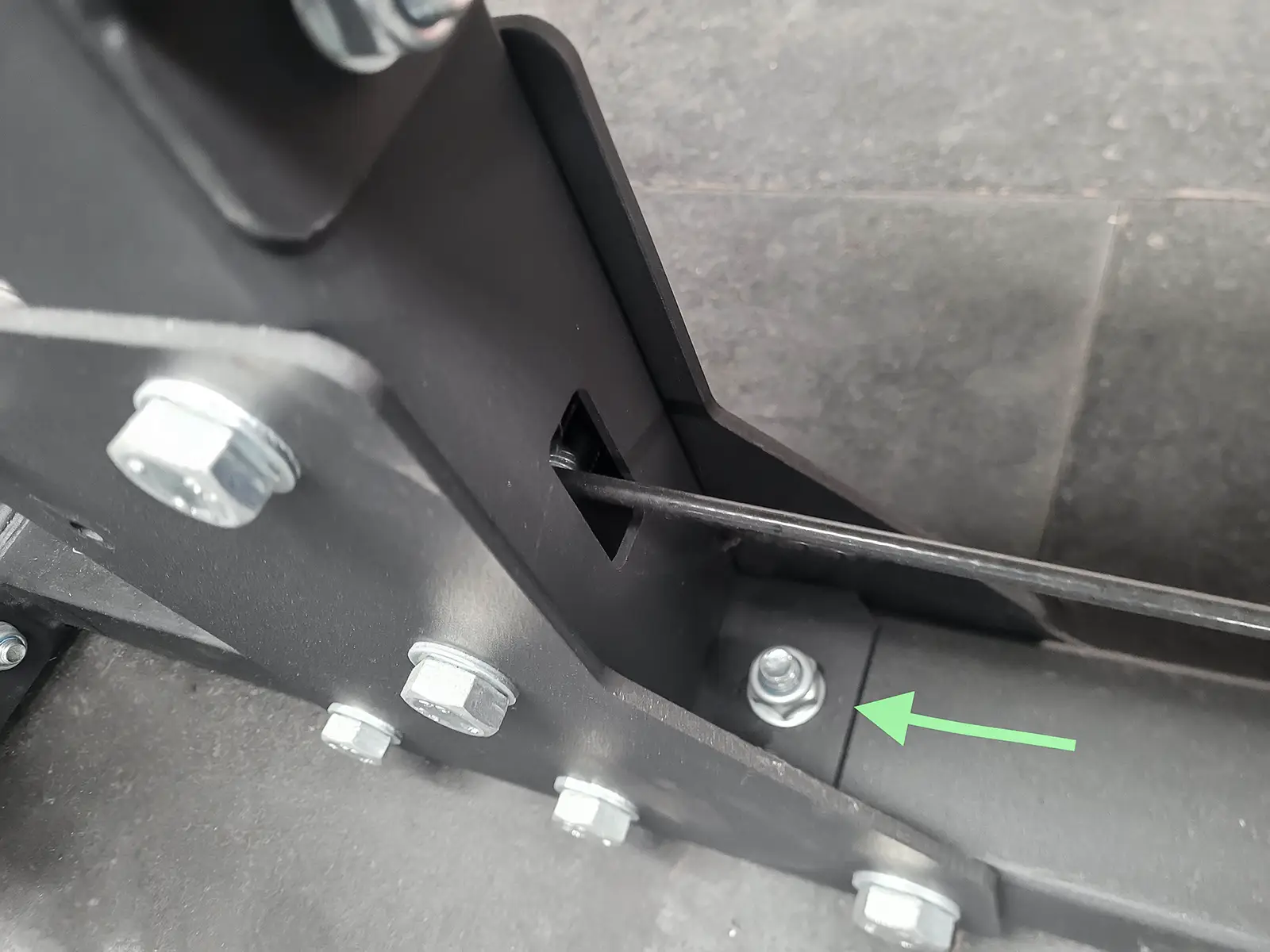 Close-up of a mechanical hinge on a lat pulldown machine, with multiple bolts attached to a metal frame. A green arrow points to a specific bolt, highlighting its placement. The background consists of a dark tiled surface.