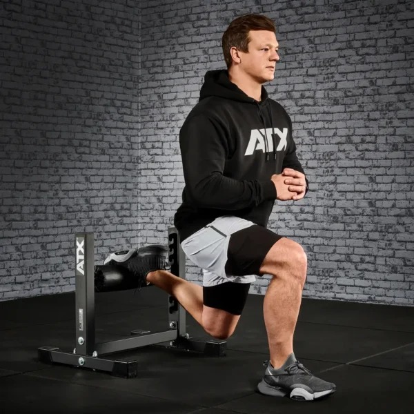 In a gym setting with a gray brick wall backdrop, someone in a black hoodie and shorts uses an ATX® Split Squat Stand to perform split squats, emphasizing balance and strength with one foot on the stand.