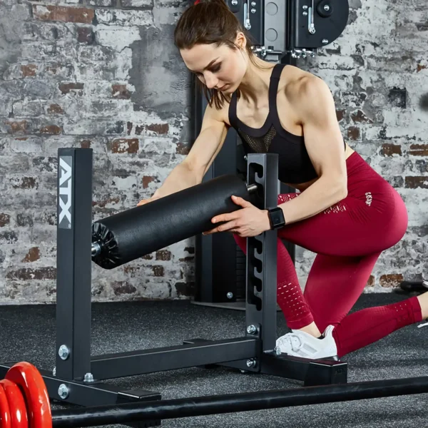 A woman in a black tank top and red leggings sets up an ATX® Split Squat Stand at a fitness center, kneeling on a mat to secure the padded roller to the metal frame against a brick wall background.