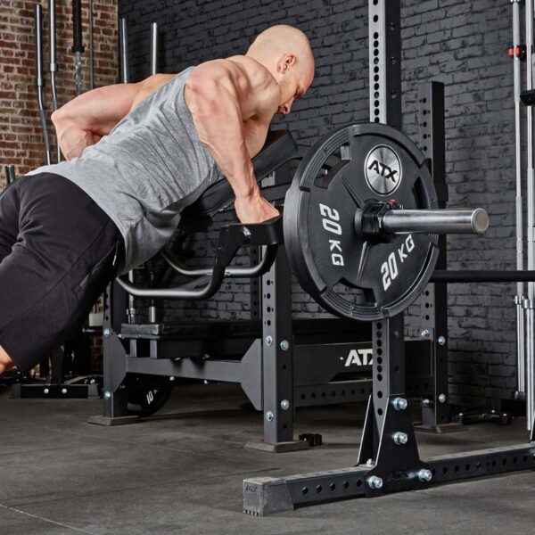 Wearing a gray tank top and black shorts, an individual uses a barbell with 20 kg plates to lift weights in a gym surrounded by black equipment and brick walls, utilizing the ATX® The Bridge Supporting Pad.