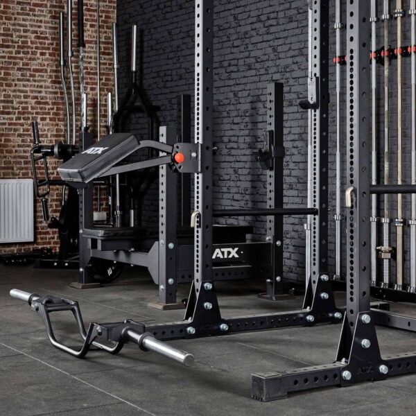 A gym with an industrial look features equipment like a power rack, weight bench, barbell with weights, and racks of weight plates. The space is accented by brick walls and dark flooring. The ATX® The Bridge Supporting Pad is part of this setup.