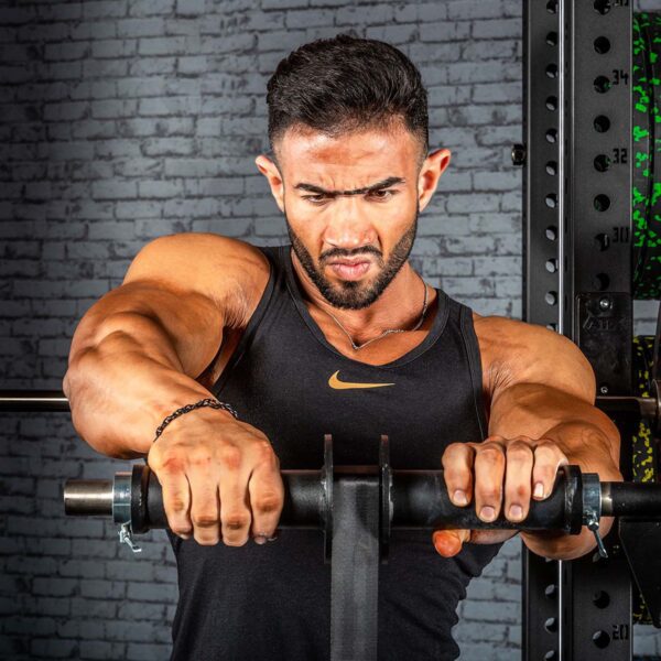 In a gym against a dark brick wall, a muscular man in a black tank top intensely grips the ATX® Forearm Training Set barbell, showcasing his strength and focus.