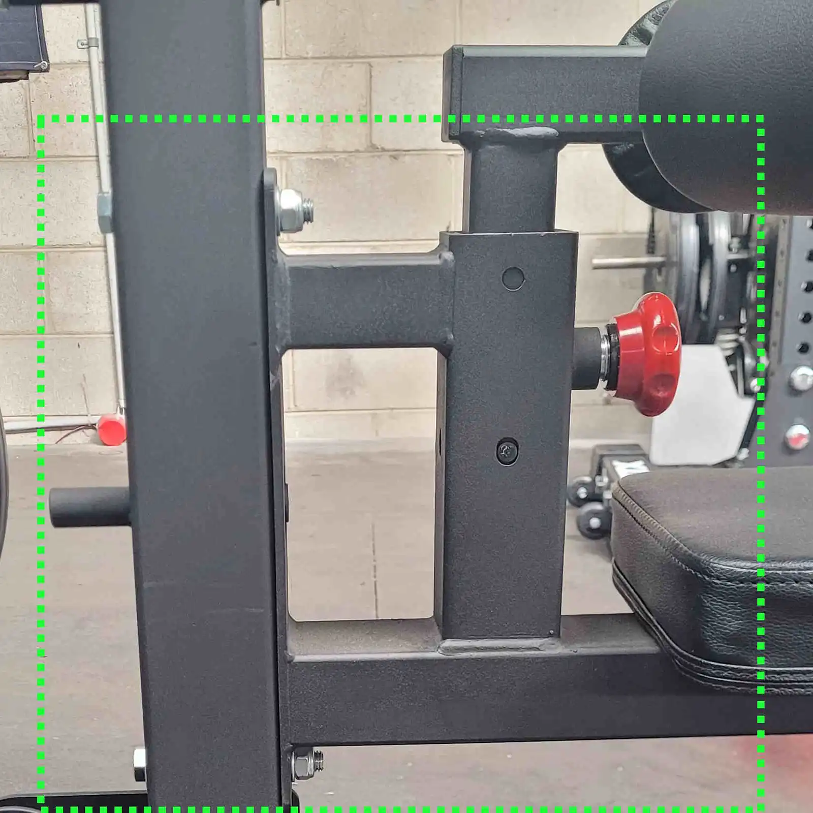 Close-up of gym equipment focusing on metal frames with a bright red adjustment knob and padded seats, highlighting the Lat Pulldown machine. The background features a concrete wall, exercise equipment, and a barbell.