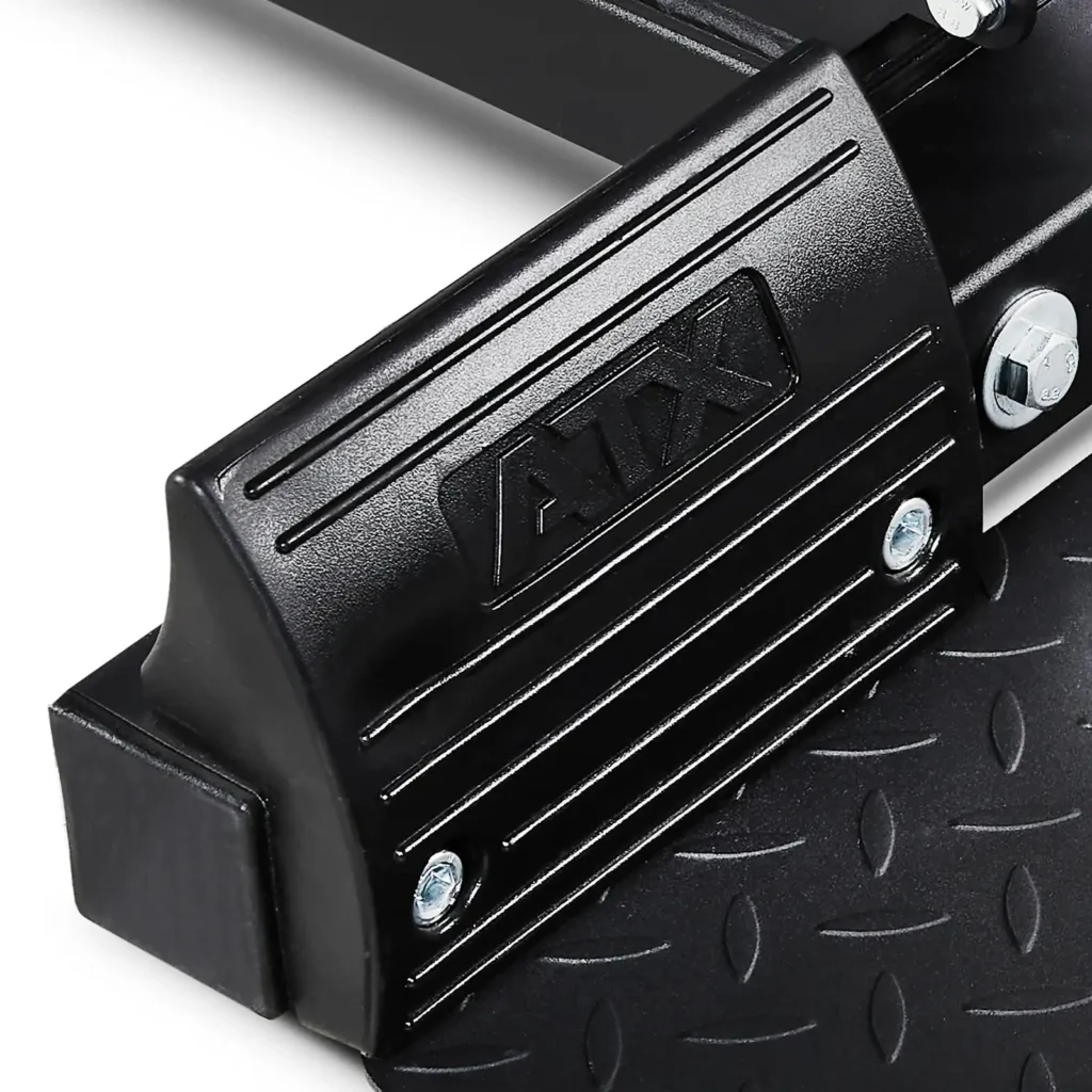 Close-up of a black metal footplate with textured grip, featuring the brand name ATX embossed in a raised design. The background reveals parts of a Lat Pulldown and Row Machine, showcasing screws and bolts intricately arranged.