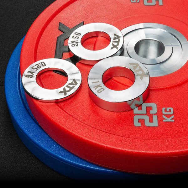 On a black surface are stacked red and blue weight plates, topped with three small silver ATX® Olympic Fractional Plate Pairs labeled 0.25 kg and 0.5 kg.