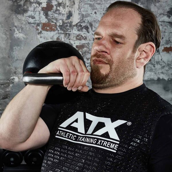 In a gym with a weathered brick wall and exercise gear, someone wearing an ATX® Grip Shirt lifts a kettlebell.