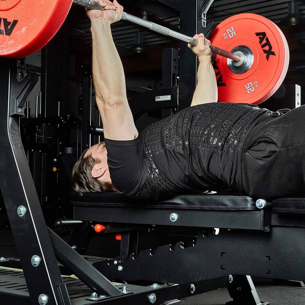A person in a black ATX® Grip Shirt and pants is bench pressing a barbell with red weight plates in a gym, emphasizing arm muscles and strength training.