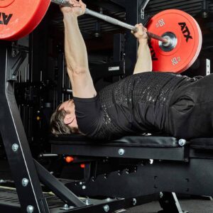 A person in a black ATX® Grip Shirt and pants is bench pressing a barbell with red weight plates in a gym, emphasizing arm muscles and strength training.