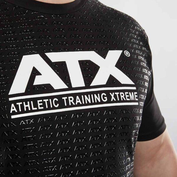 Close-up of a person wearing a black ATX® Grip Shirt featuring a repeating ATX pattern. A larger ATX logo and Athletic Training Xtreme text are prominently printed in white on the chest, highlighting style and strength.