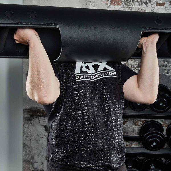 Someone wearing an ATX® Grip Shirt is lifting a heavy black weight bar. Only their arms and upper body are visible against a backdrop of gym equipment and a brick wall.