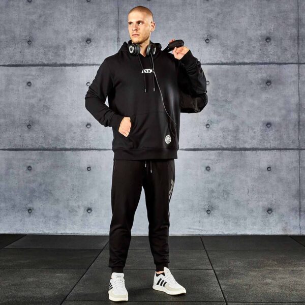 A man in an ATX® Black Hoodie and joggers stands in a gym, holding a black gym bag. He wears headphones around his neck and white sneakers, positioned against a concrete wall.