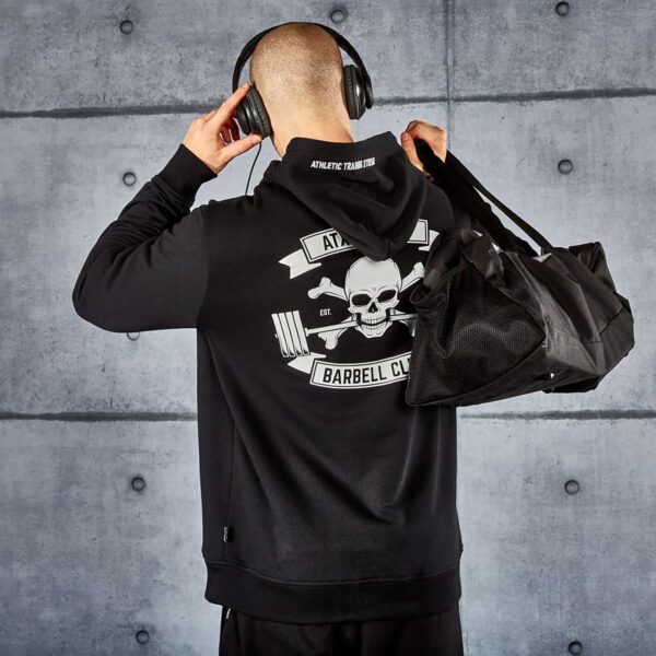 A person wearing the ATX® Black Hoodie featuring a skull and crossbones on the back stands against a textured gray wall. They carry a black gym bag and wear headphones, looking away from the camera.