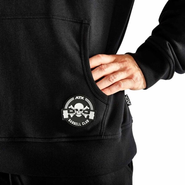 A person is wearing an ATX® Black Hoodie featuring the ATX Barbell Club logo with a skull and barbells. Their right hand rests on the hip pocket, and the hoodie tag is visible on the side.