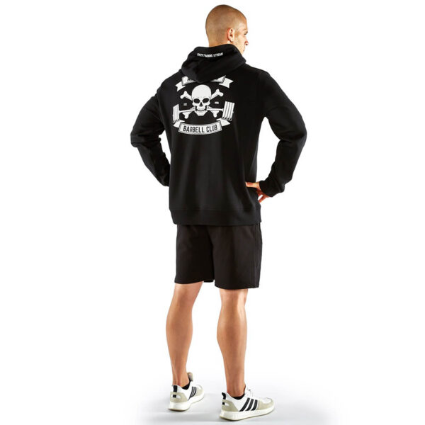 A man wearing an ATX® Black Hoodie with a skull and crossbones design and Barbell Club text on the back stands with his back to the camera, paired with black shorts and white sneakers featuring black stripes.