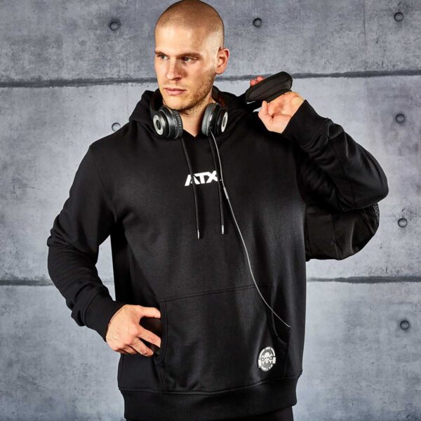 A man wearing an ATX® Black Hoodie stands against a concrete wall with headphones around his neck, holding a black device over his shoulder and looking off to the side.