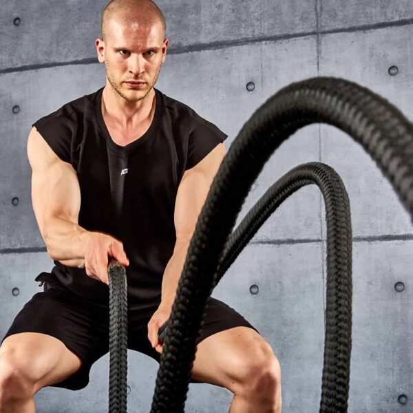 Wearing an ATX® Muscle Shirt and shorts, a person exercises intensively with battle ropes in a gym setting, with a gray concrete wall in the background, reflecting determination on their face.