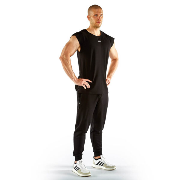A man in an ATX® Muscle Shirt, sleeveless and black, paired with black jogging pants, stands with his hands on hips. He sports white sneakers featuring black stripes against a plain white backdrop.