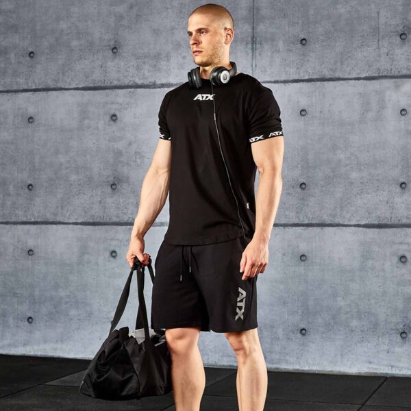 In a gym, someone stands with a duffel bag, wearing ATX® Black Shorts and a black T-shirt branded with ATX. Headphones rest around their neck against a backdrop of a concrete wall.