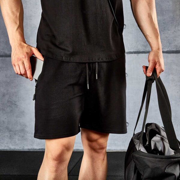 A person in black workout attire, featuring a pair of ATX® Black Shorts and a matching t-shirt, holds a gym bag while standing against a gray concrete wall with their face not visible in the image.