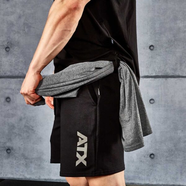 A person in a black shirt and ATX® Black Shorts, with ATX on the side, ties a gray shirt around their waist. A gray concrete wall with visible seams is in the background.