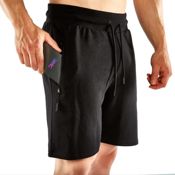 A man wearing ATX® Black Shorts with drawstrings and zippered pockets holds a smartphone partly tucked into one of the pockets.