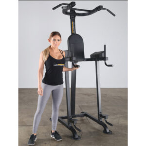 A woman in athletic attire stands beside a Powertec Basic Trainer in an empty room. Shes wearing a black tank top, gray leggings, and black shoes, smiling slightly. The sleek equipment is designed for pull-ups and dips.