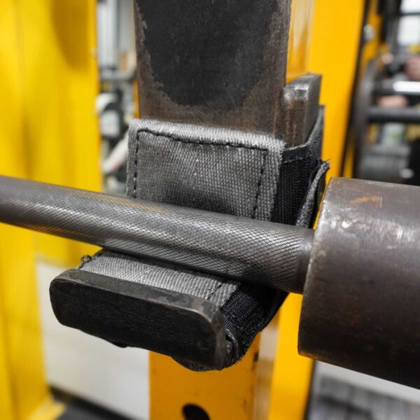 A close-up of the Spud Inc 5R Barbell Shield resting securely on a safety catch in a bright yellow squat rack highlights the equipments texture and material.