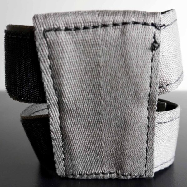 A close-up of the Spud Inc 5R Barbell Shield shows a gray fabric strap with black stitching and a black elastic section, folded to display its texture. Its set on a smooth, dark surface.