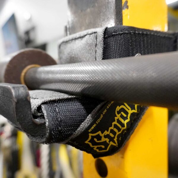 Close-up of a Spud Inc 5R Barbell Shields padded strap holding a barbell, secured on a yellow metal frame. The strap displays the logo and spudincstraps.com. Slightly out-of-focus gym equipment is seen in the background.