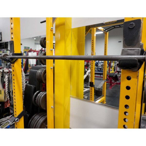 A gym featuring a yellow power rack with weight plates stacked on the side and a Spud Inc 5R Barbell Shield resting on hooks. Exercise equipment and mirrors are visible in the background.