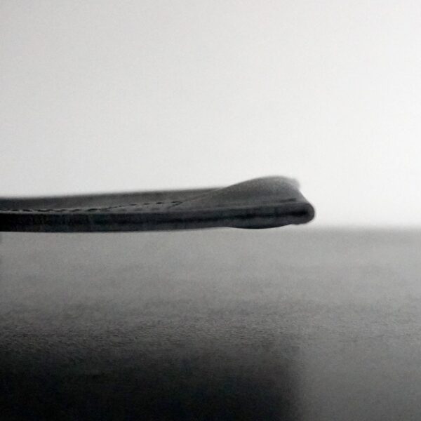A close-up of a thin, flat Spud Inc The Gripazz on a dark surface against a light background. It has a slightly raised, curved edge and appears to be made of gray material, possibly leather or fabric.