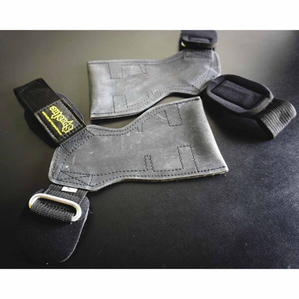 A close-up of the Spud Inc The Gripazz, two black lifting straps with padded supports and metal rings on a dark surface, featuring yellow stitching and a weightlifting logo.