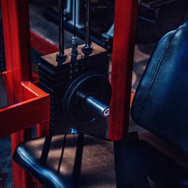 A close-up of a gym machine showcases a sleek black leather seat and red metal frame, with weight plates easily adjusted using the GymPin 2 Original, and numbers visible on the dimly lit weight stack.