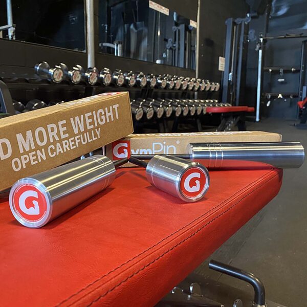 Cylindrical weights with red and white logos rest on a red bench alongside an open box labeled ADD MORE WEIGHT, showcasing a new GymPin 2 Original. In the background, a gleaming rack of silver dumbbells reflects off the mirror.