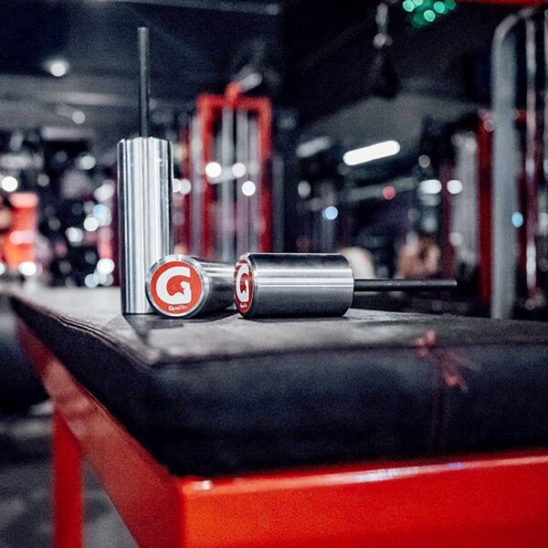Close-up of two metallic GymPin 2 Original scent diffusers with red logo caps on a gym bench, set against gym equipment and blurred lights, creating a modern, energetic atmosphere.