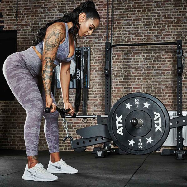 A woman in workout gear performs a bent-over row with the ATX® Belt Squat Option in a gym. She focuses amid fitness equipment, standing on a black rubber floor with brick walls behind her.