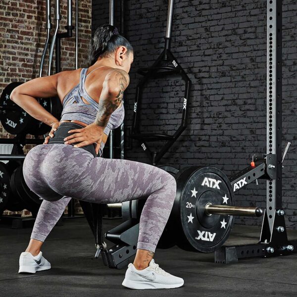 A person in athletic wear performs a squat using the ATX® Belt Squat Option in a gym. Theyre surrounded by workout equipment, such as weight plates and racks, against a brick wall.