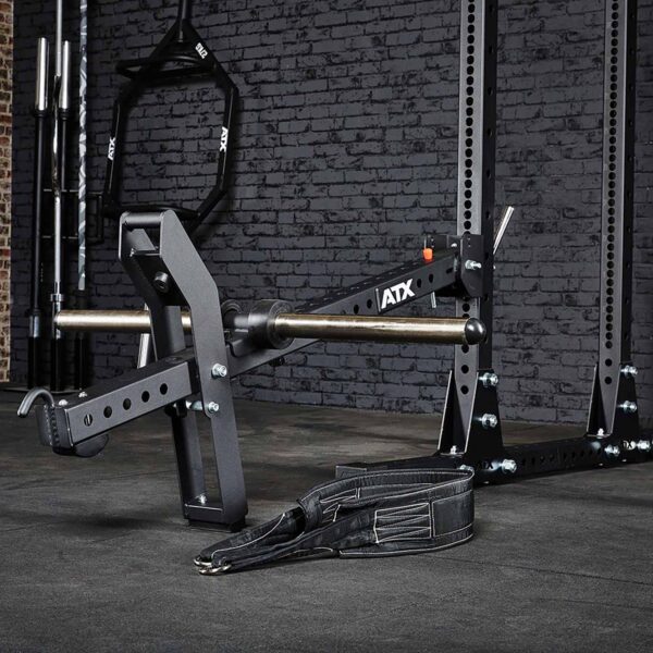 The ATX® Belt Squat Option features a weightlifting machine with a barbell, black padded straps, and adjustable metal arms. Designed for strength training against a dark brick wall, it includes secure bolts and a sturdy frame.