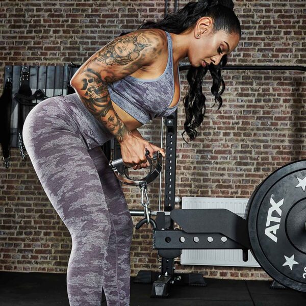 A tattooed woman in a gray camo workout outfit performs a bent-over barbell row with the ATX® Belt Squat Option in a gym, backed by a brick wall and various equipment.