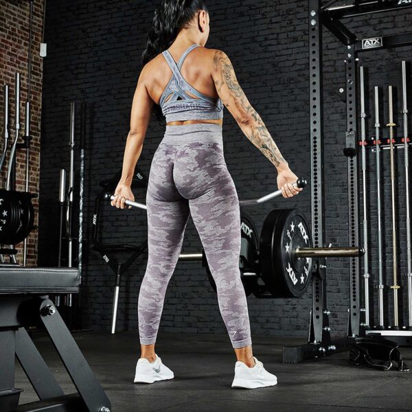 A person with long hair, wearing a sports bra and camouflage leggings, is lifting a barbell in a gym equipped with an ATX® Belt Squat Option. The space features brick walls and various workout gear like racks and benches.