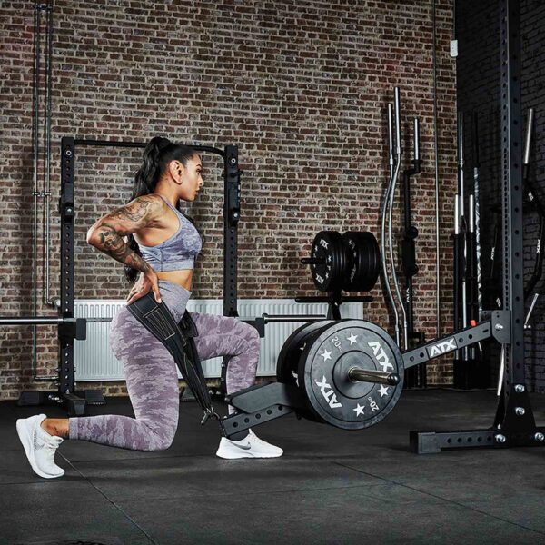A woman in camo workout gear lunges with a resistance band at the gym. The backdrop showcases brick walls, weights, racks, and an ATX® Belt Squat Option.
